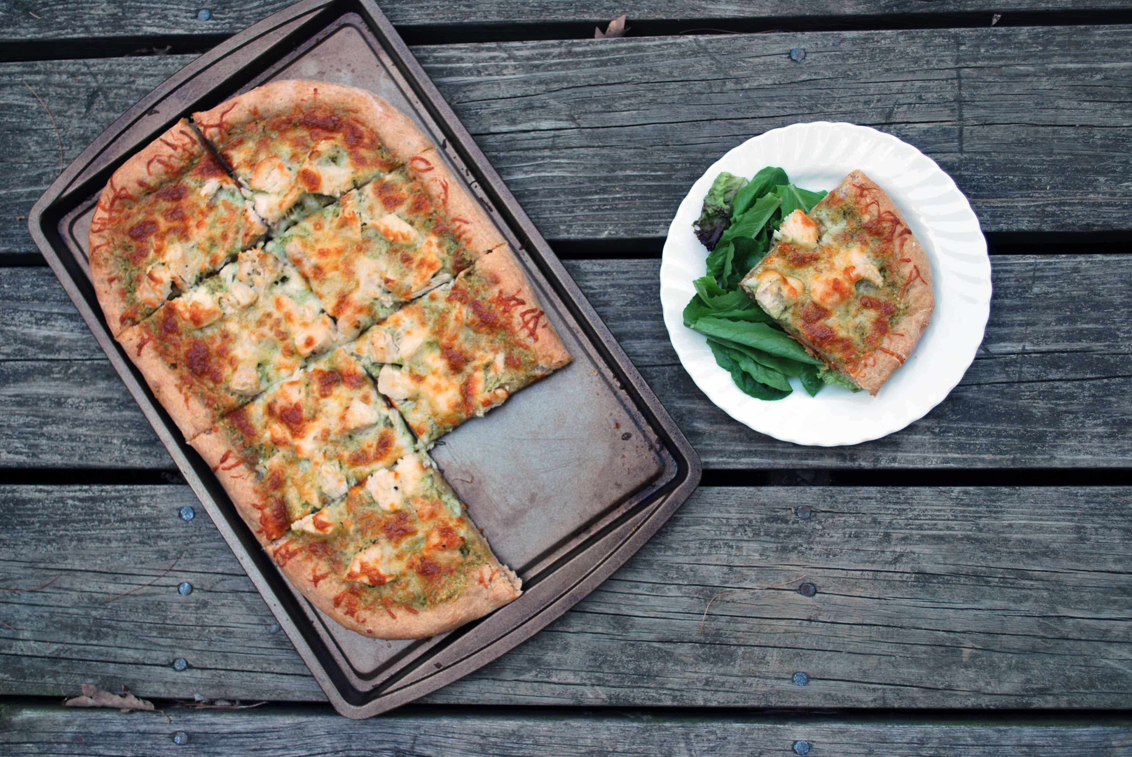 PestoChickenPizzaOHWS a slice of Pesto Chicken Pizza on a white plate next to a side salad and a sheet pan of pizza