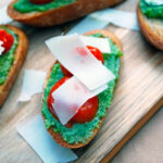 Pesto and Oven Roasted Tomato Bruschetta on a wooden cutting board topped with shaved parmesan cheese.