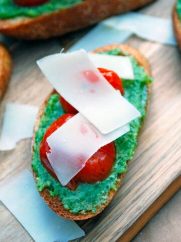 Pesto and Oven Roasted Tomato Bruschetta on a wooden cutting board topped with shaved parmesan cheese.