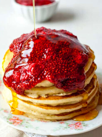 syrup being poured on a stack of pancakes topped with chia jam.