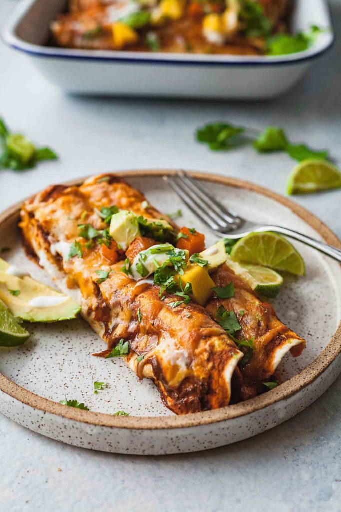 two black bean and butternut squash enchiladas on a rustic plate with avocado, limes, and a fork