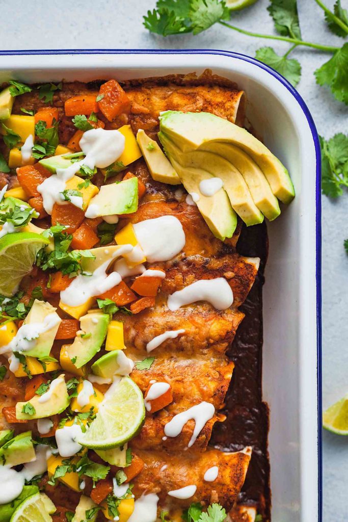 a pan of butternut squash enchiladas topped with avocado, mango, lime, and cilantro