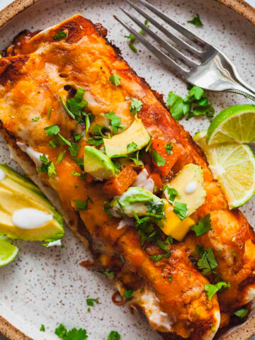 a speckled plate with two butternut squash enchiladas, limes, and avocado.