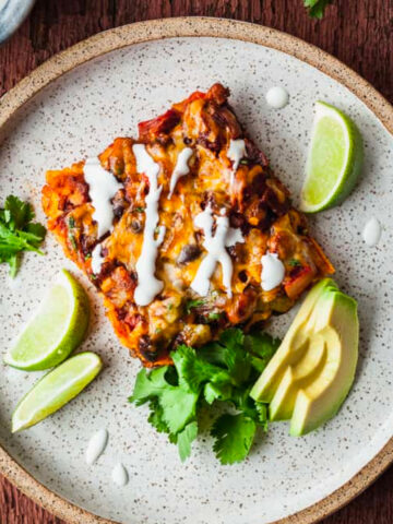 a slice of stacked veggie enchiladas on a rustic plate with cilantro, limes, and avocado.