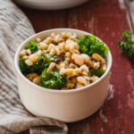 close up image of small white bowl with barley salad on red picnic table with bigger bowl of barley salad in background.