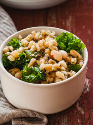 close up image of small white bowl with barley salad on red picnic table with bigger bowl of barley salad in background.
