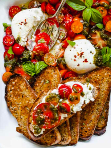 white casserole dish with burrata with tomatoes pesto and toast with a metal spoon.