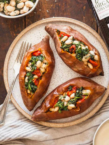 Vegan stuffed sweet potatoes with spinach and white beans on a speckled plate with a fork, against a wood background with a grey striped towel and bowl of tahini sauce.