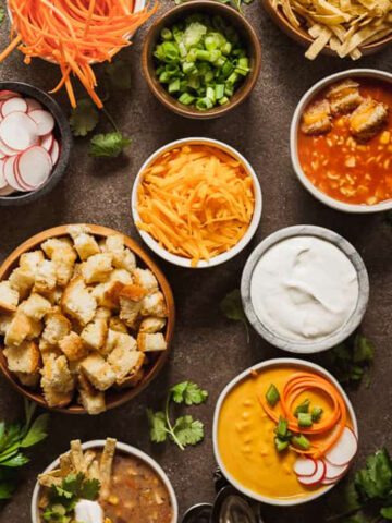 many bowls filled with soup and toppings for soup such as croutons, cheese, radishes, and sour cream.