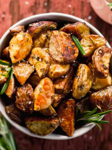 a bowl of oven roasted potatoes with rosemary on a red wood background with rosemary sprigs and salt.