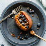 a sweet potato stuffed with blueberries, pecans, and chia seeds, drizzled with almond butter and maple syrup on a blue plate with a fork and wood spoon