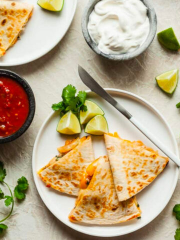 two white plates with chipotle veggie quesadillas, a knife, limes, and bowls of salsa and sour cream.