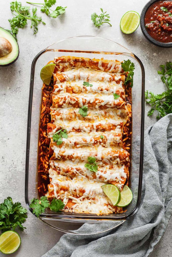 a pan of cheese enchiladas with a towel, bowl of salsa, limes, and cilantro