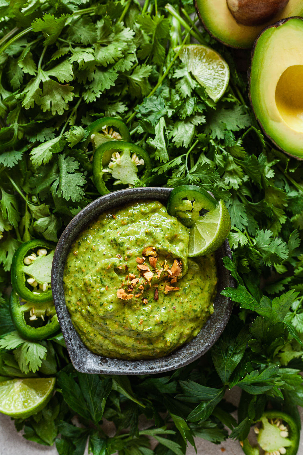 creamy avocado sauce in a small bowl surrounded by herbs, avocados, and tortilla chips