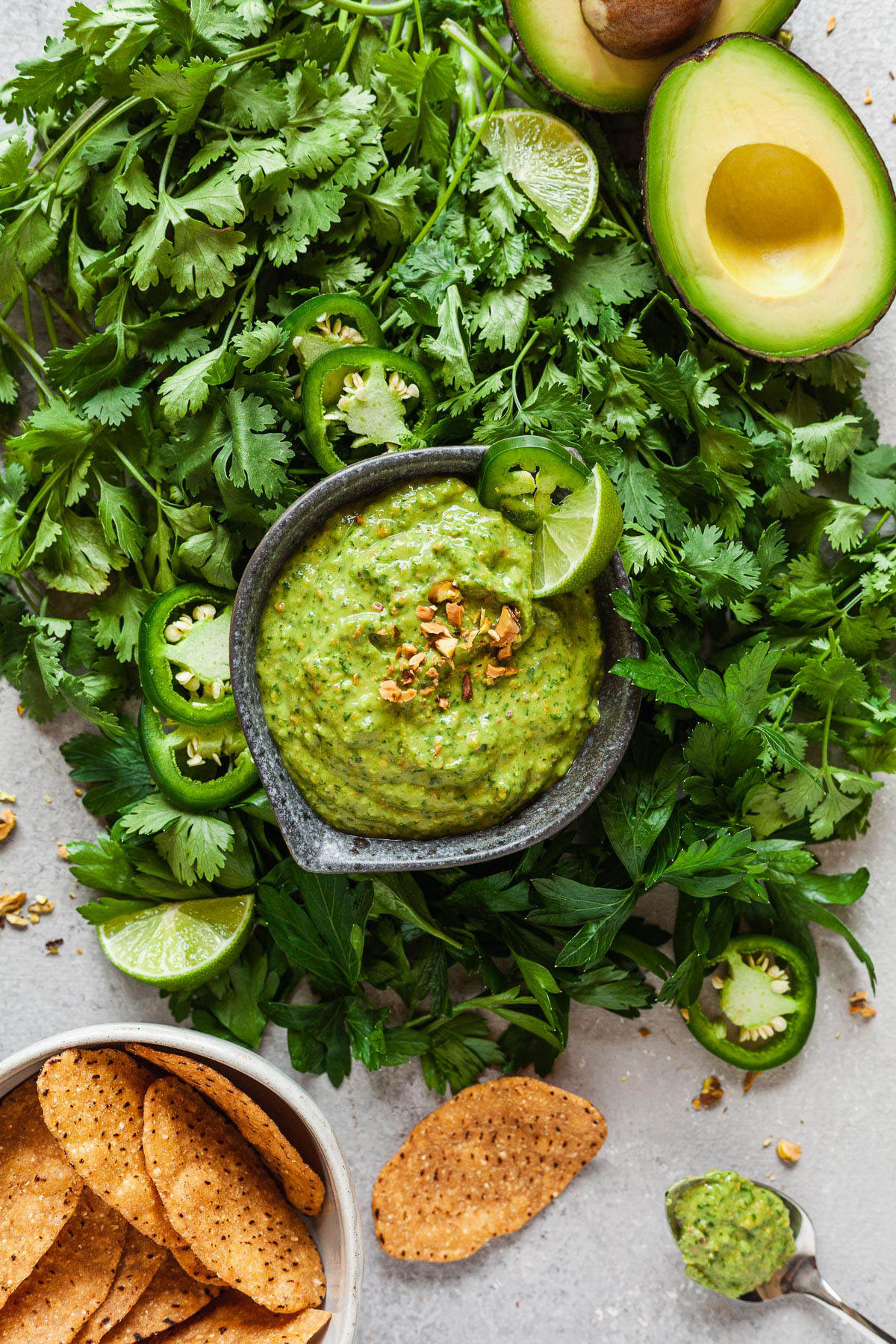 green avocado sauce in a small bowl surrounded by herbs, avocados, and tortilla chips