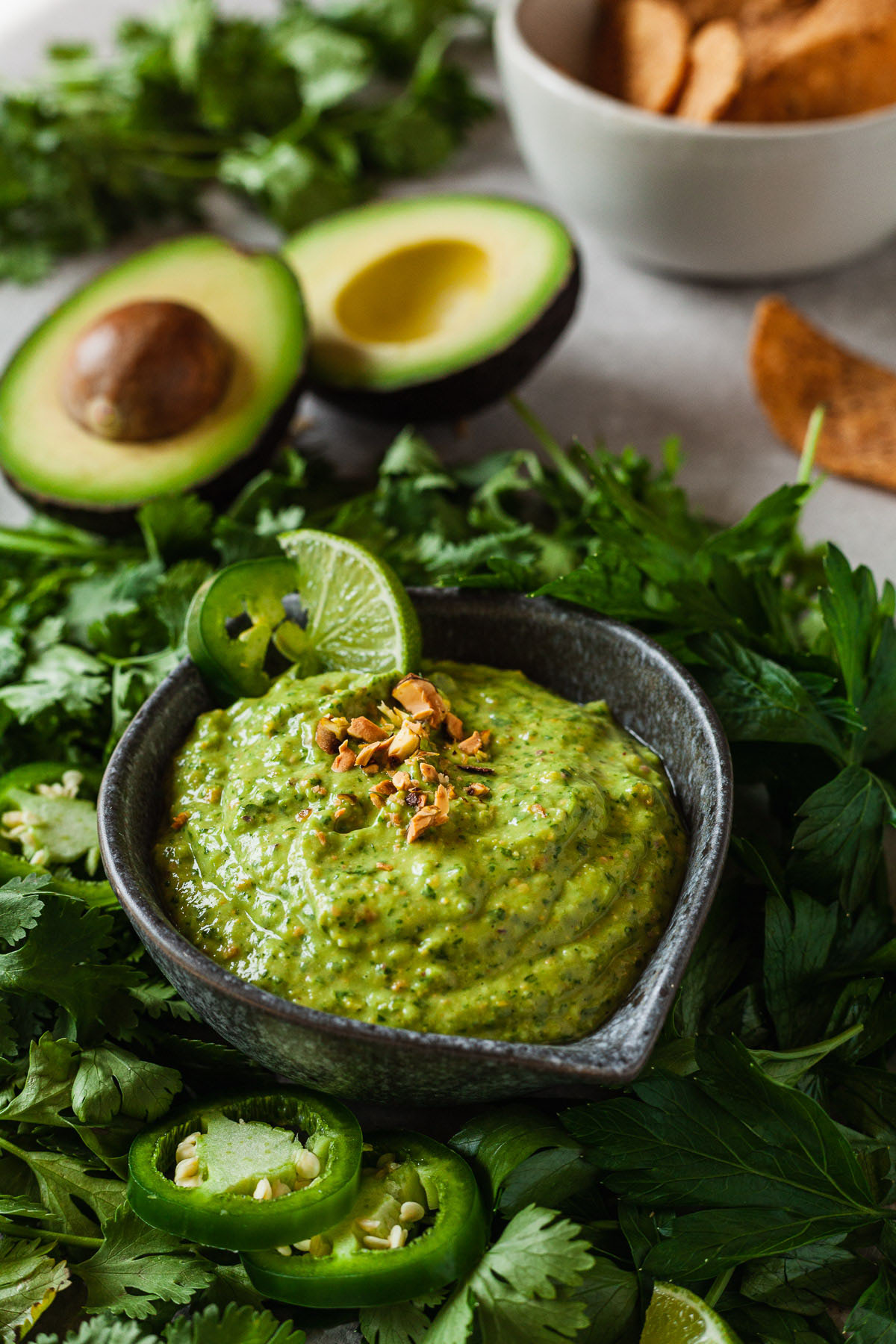 green avocado sauce in a small bowl surrounded by herbs, avocados, and tortilla chips.