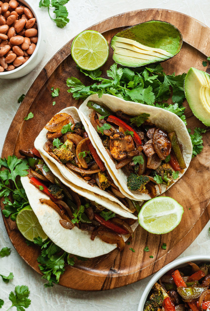 three vegan fajitas on a platter with limes, avocado, and cilantro