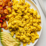 scrambled tofu eggs on a white plate with home fries and avocado slices
