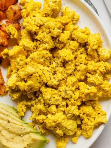scrambled tofu eggs on a white plate with home fries and avocado slices