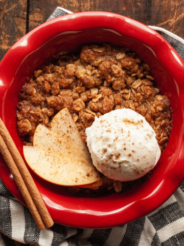 individual apple crisp in a red pie plate with apple, cinnamon stick, and ice cream
