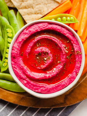 vibrant pink beetroot hummus in a bowl on a platter of crackers, peas, and carrots