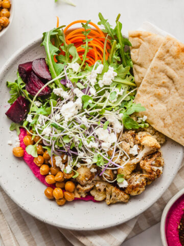 a close up image of a hummus bowl filled with chickpeas, beet hummus microgreens, pita bread, and more