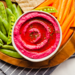 vibrant pink beetroot hummus in a bowl on a platter of crackers, peas, and carrots
