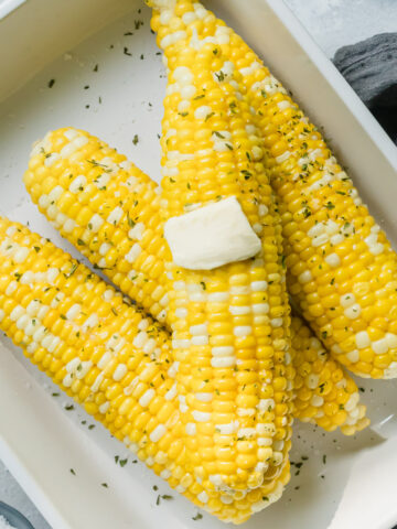 ears of corn in a white baking dish topped with a pat of butter