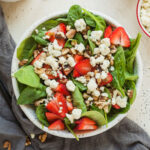 strawberry spinach salad in a bowl next to a small bowl of strawberries, a small bowl of gorgonzola cheese, and a little container of poppy seed dressing