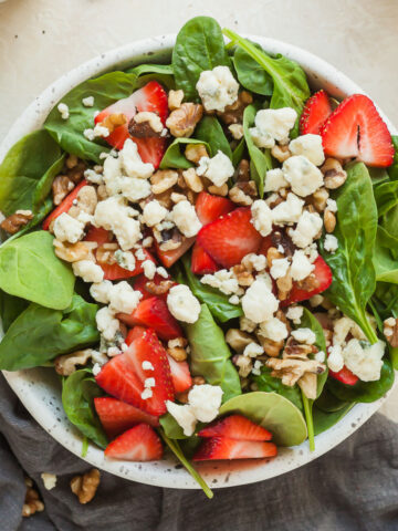 strawberry spinach salad in a bowl next to a small bowl of strawberries, a small bowl of gorgonzola cheese, and a little container of poppy seed dressing