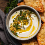 whipped feta dip topped with olive oil, herbs, and pistachio served in a bowl on a plate next to crostini