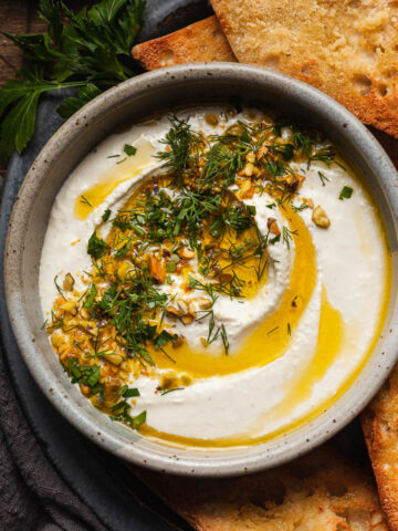 whipped feta dip topped with olive oil, herbs, and pistachio served in a bowl on a plate next to crostini