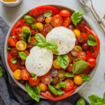burrata caprese on a round plate next to a gray linen napkin, a little jug of olive oil, a bowl of salt, and a spoon