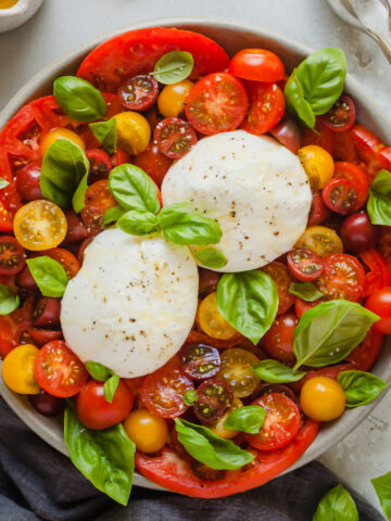 burrata caprese on a round plate next to a gray linen napkin, a little jug of olive oil, a bowl of salt, and a spoon