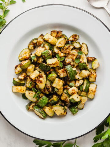 Air fryer zucchini on a round plate next to a spoon and green herbs.