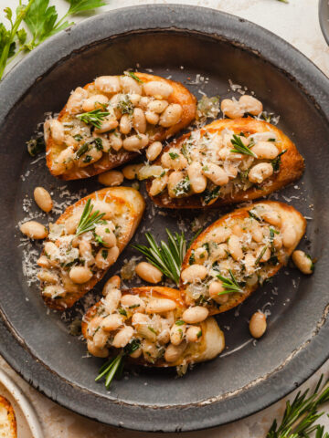 White bean crostini on a gray plate topped with fresh rosemary and parmesan cheese, next to a bunch of parsley and a sprig of rosemary.