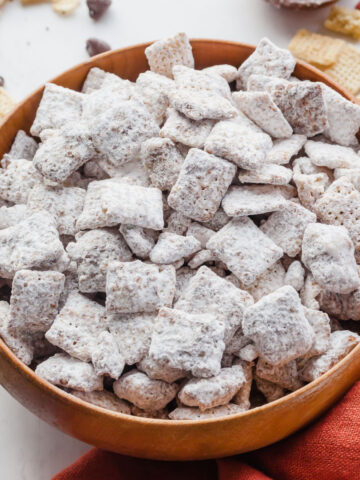 Puppy chow in a wooden bowl surrounded by rice chex and chocolate chips.