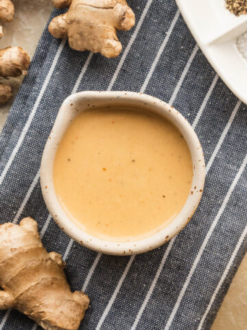 Ginger tahini dressing in a small bowl on top of a blue and white striped napkin next to lemons and fresh ginger.