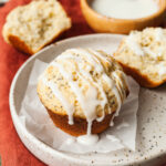 Lemon poppy seed muffins on a speckled plate on a red napkin next to a spoon of poppy seeds and a bowl of glaze.