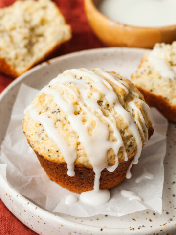 Lemon poppy seed muffins on a speckled plate on a red napkin next to a spoon of poppy seeds and a bowl of glaze.