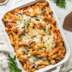 A baking dish full of meatless baked ziti on a linen striped napkin next to bunches of parsley.