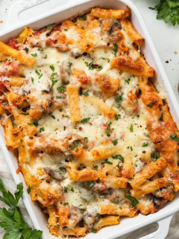 A baking dish full of meatless baked ziti on a linen striped napkin next to bunches of parsley.
