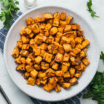 A bowl of air fryer sweet potato cubes on a striped blue napkin next to bunches of parsley.