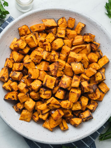 A bowl of air fryer sweet potato cubes on a striped blue napkin next to bunches of parsley.