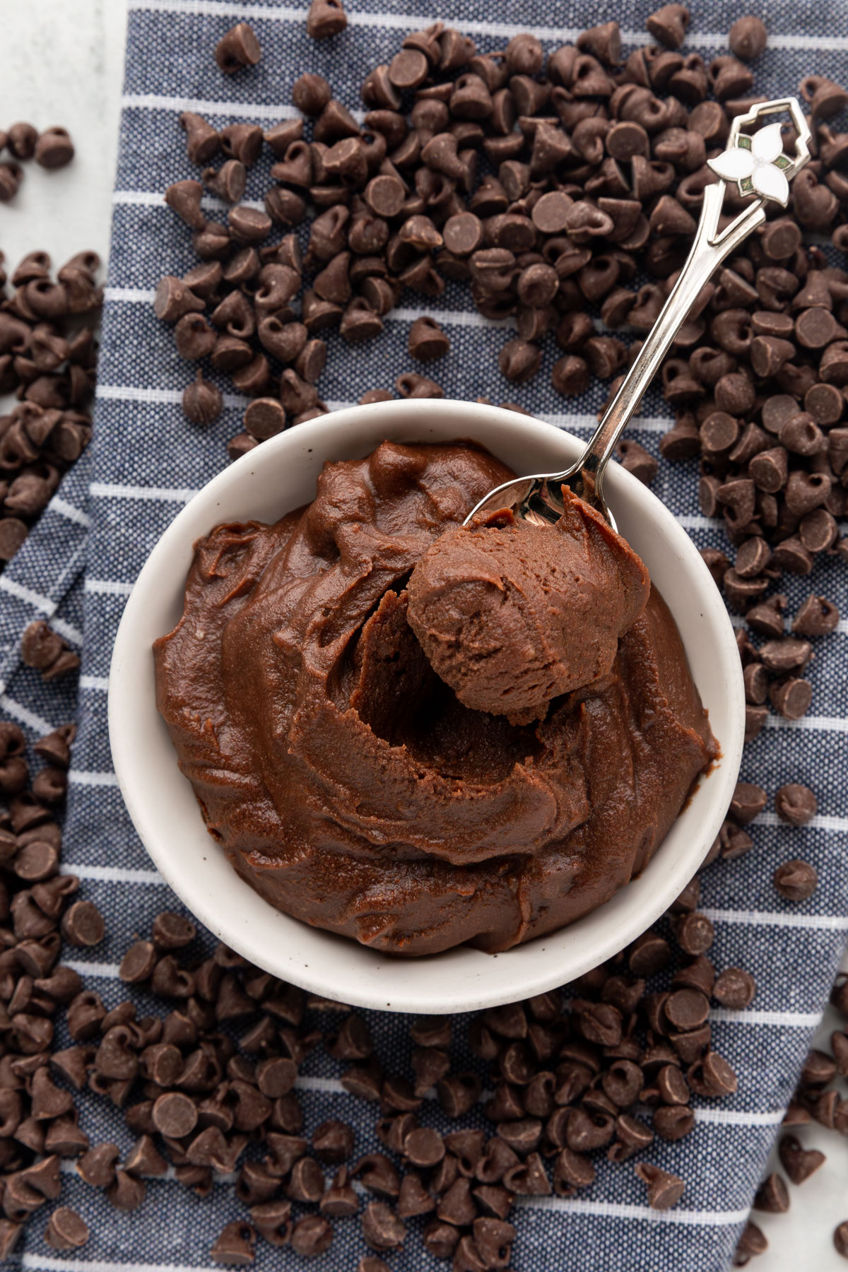 Edible brownie batter with a spoon sticking out of it on a blue and white striped napkin surrounded by chocolate chips.