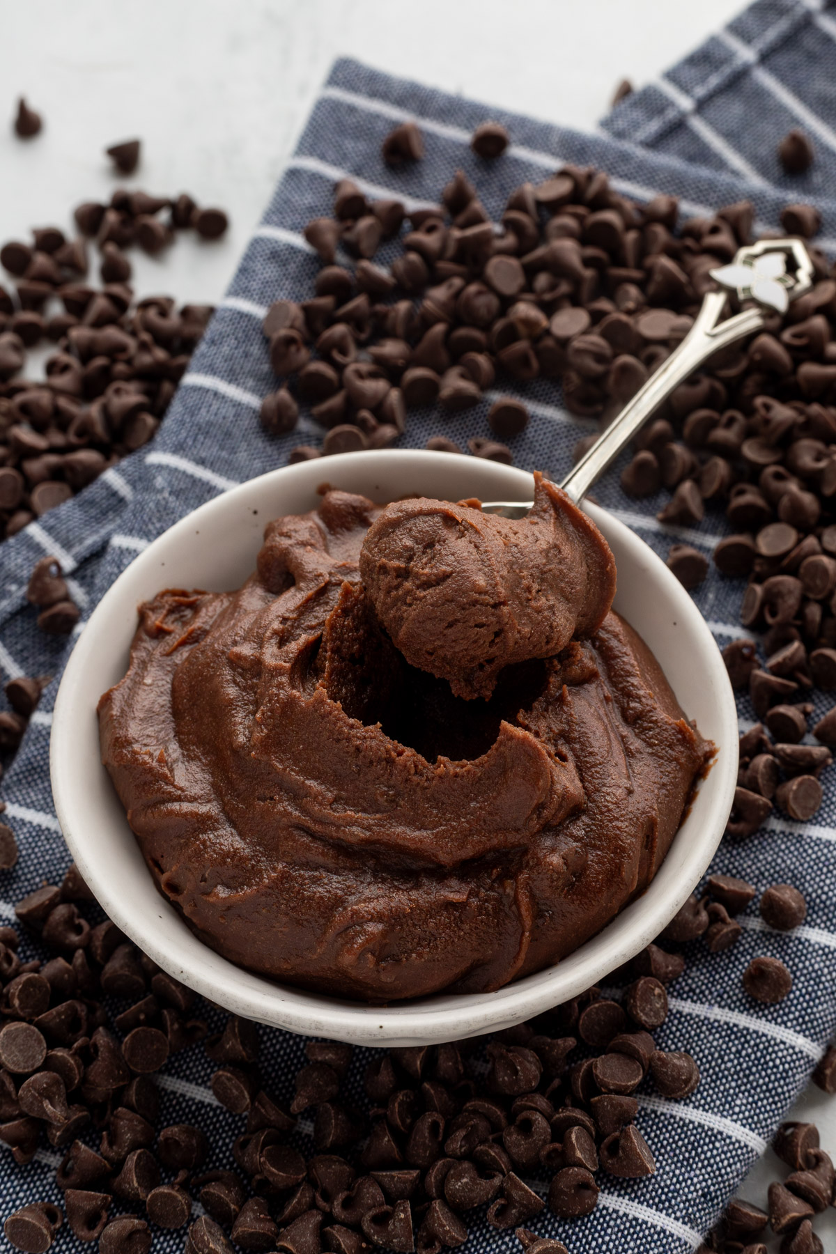 Edible brownie batter with a spoon sticking out of it on a blue and white striped napkin surrounded by chocolate chips.