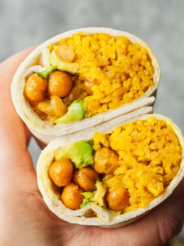 A hand holding up a An Avocado, Yellow Rice, and Roasted Chickpea Burrito cut in half.