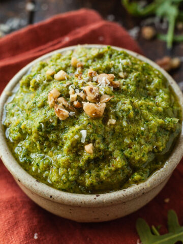 A small bowl of pesto made from arugula and walnuts topped with walnuts on top of a red napkin.