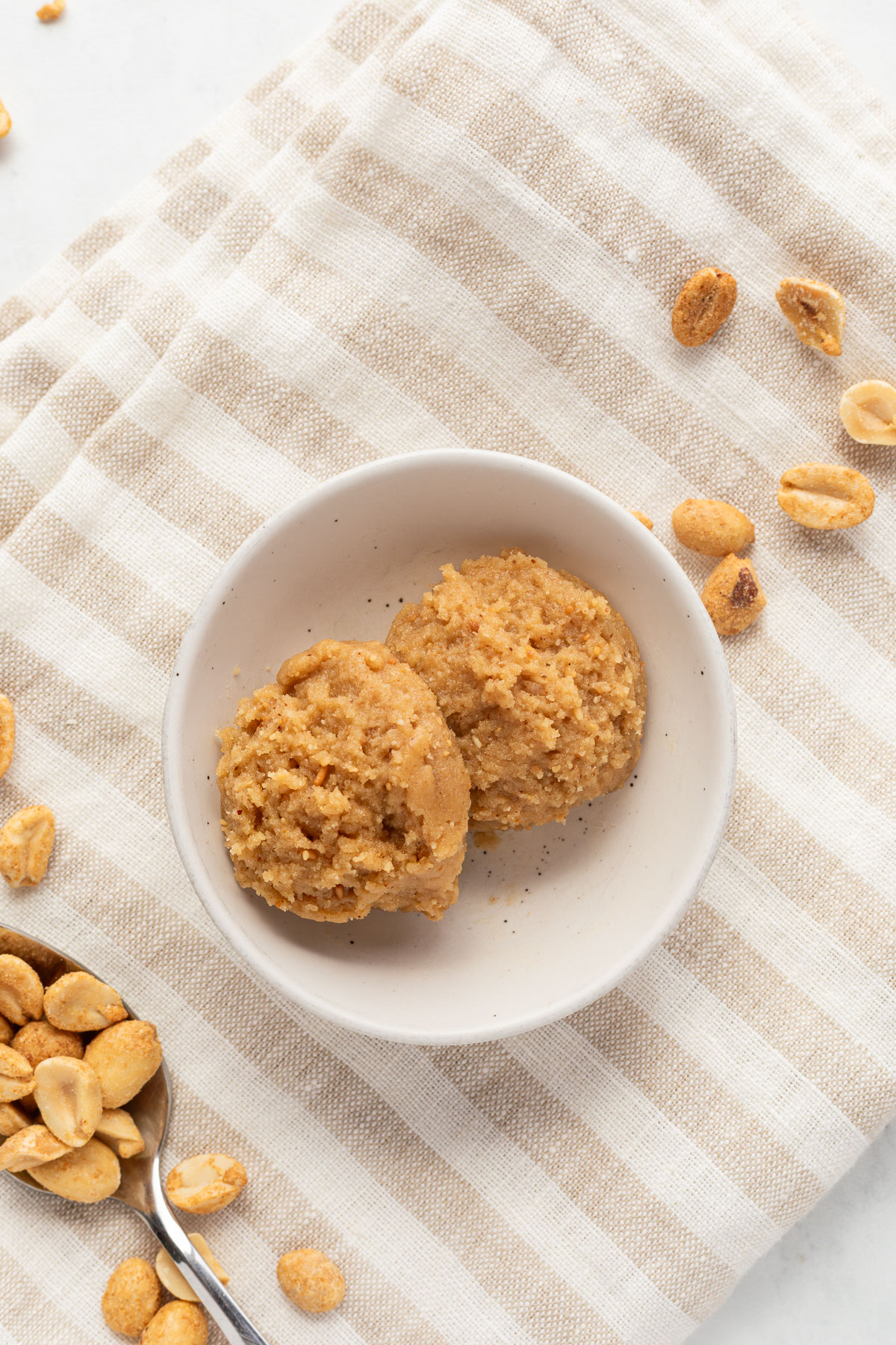 Two scoops of edible peanut butter cookie dough in a small white bowl on a striped linen napkin surrounded by peanuts.