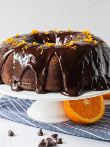 An orange chocolate cake topped with chocolate ganache and orange zest on a cake stand next to a blue and white striped napkin and orange halves.